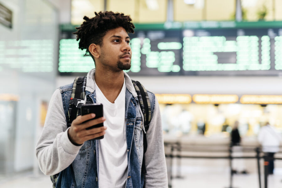 Young man inside the airport holding a mobile phone.young afro man holding a mobile phone looking at the departures of planes in an airport